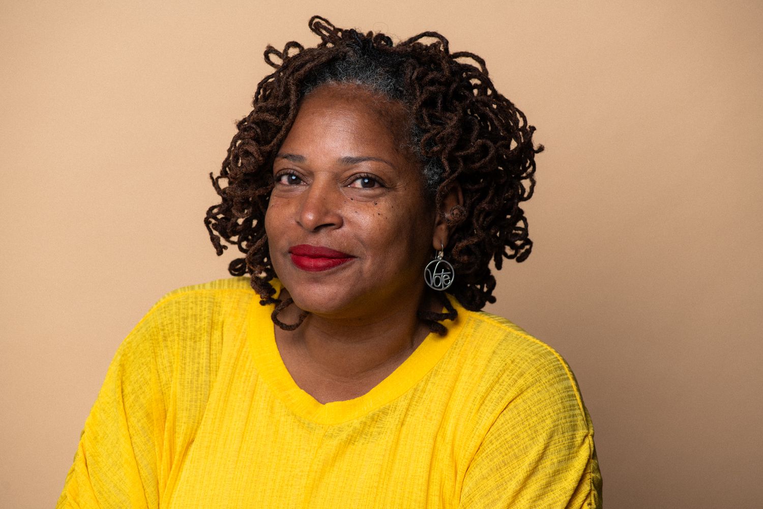 Smiling woman in yellow top with curly hair and red lips wearing teal earrings.