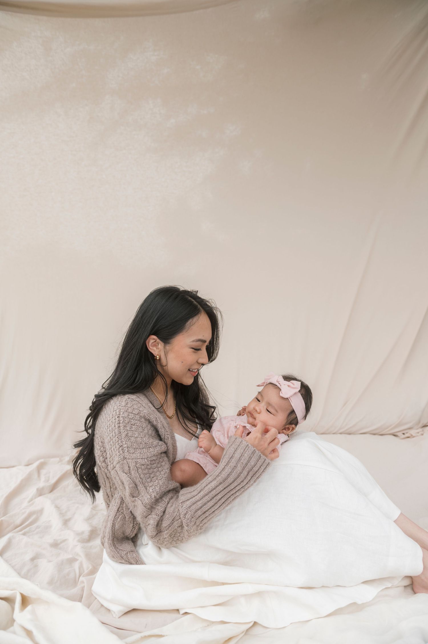 A mother in a beige knit sweater holds her newborn baby girl with a pink bow headband against a soft cream backdrop.