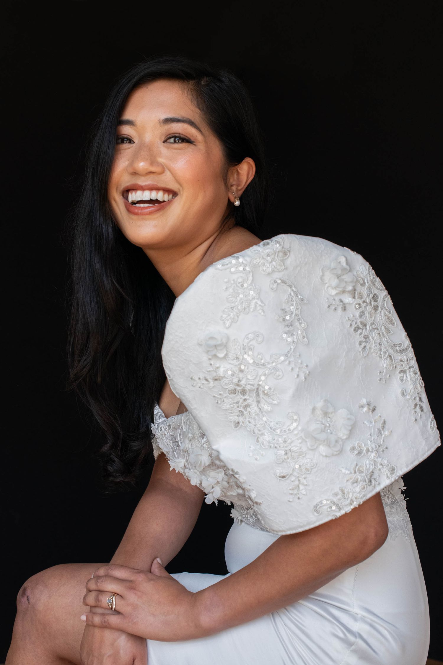 Smiling bride in white lace floral cape wedding dress poses against dark background.