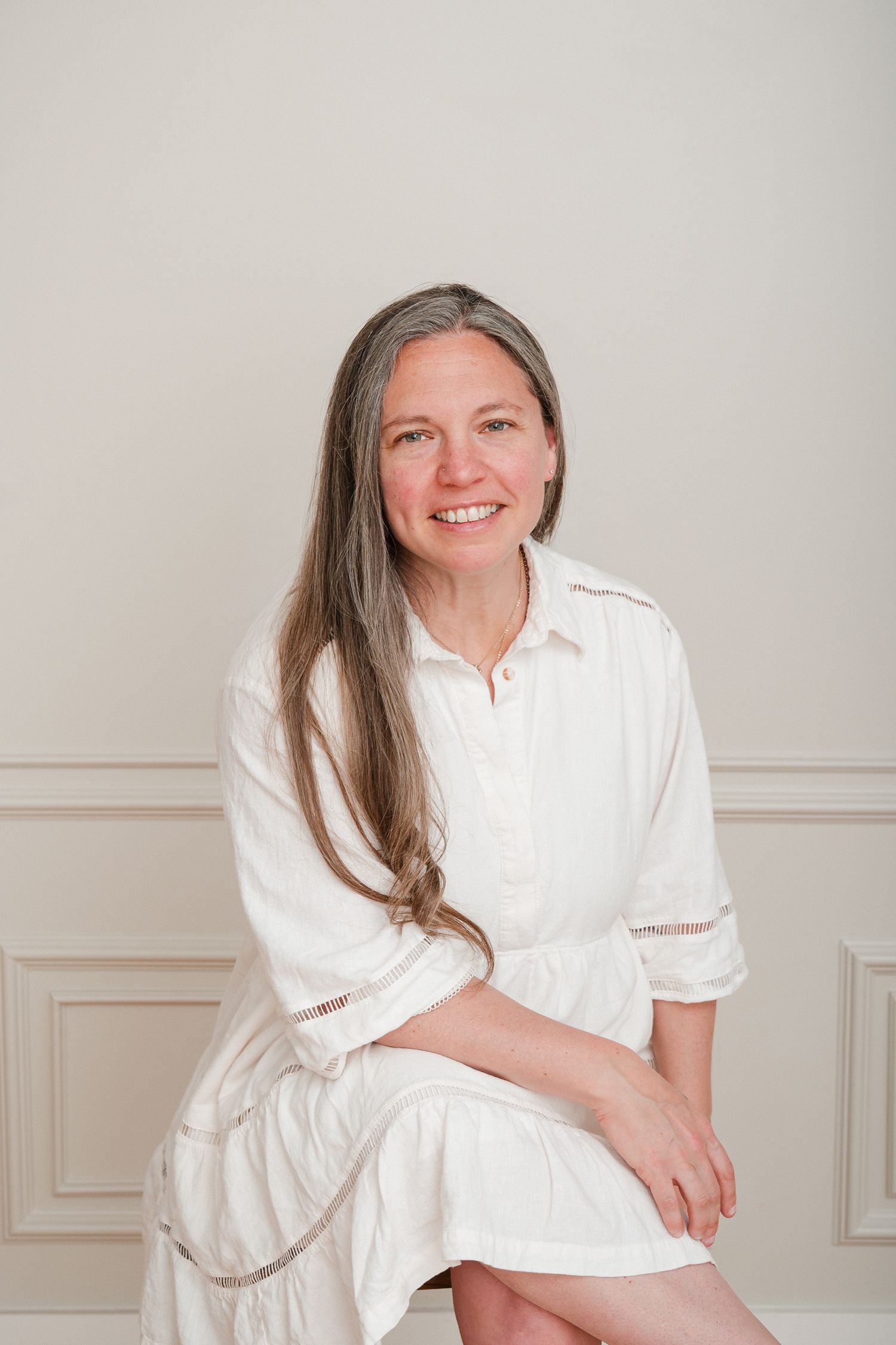 Woman in white clothing poses gracefully against a light gray wall with minimal studio backdrop.