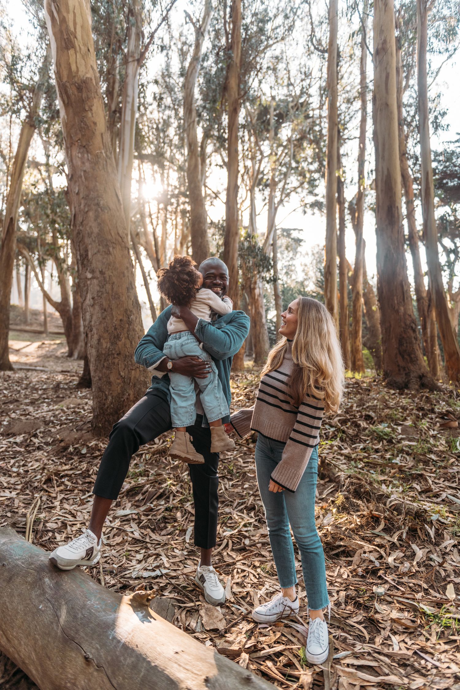 Two women and a child enjoy a sunny day in a forest, with one woman lifting the smiling child near tall trees.