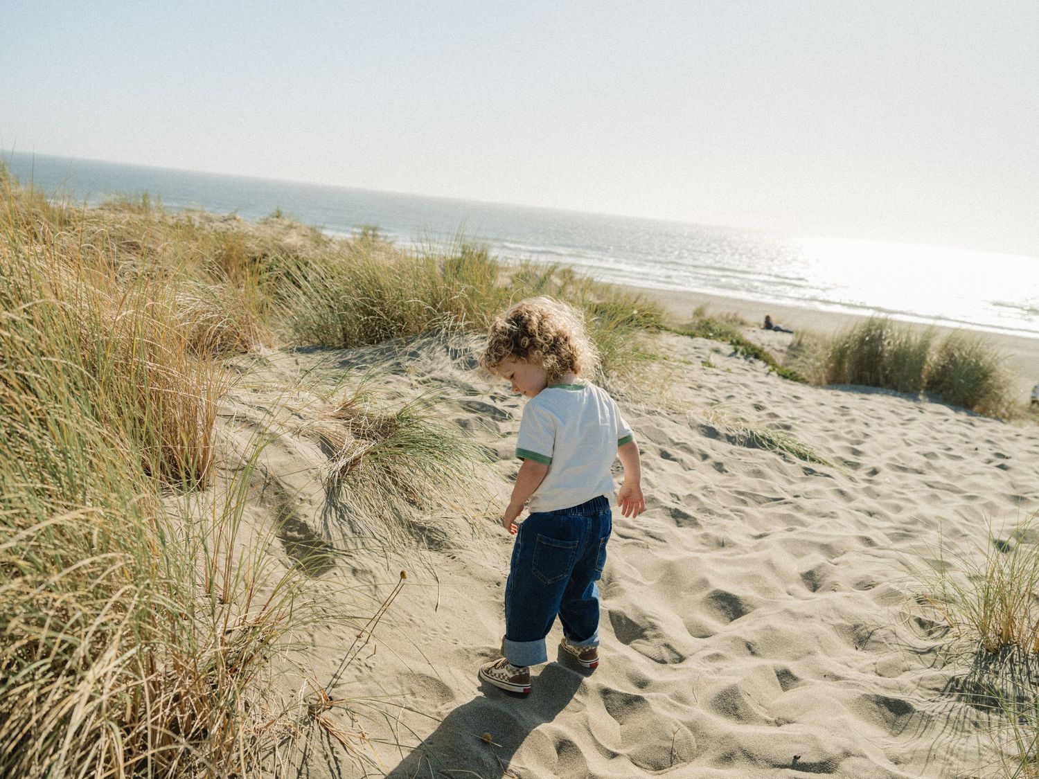A toddler with curly hair walks on sandy beach dunes with grass and ocean in the background.