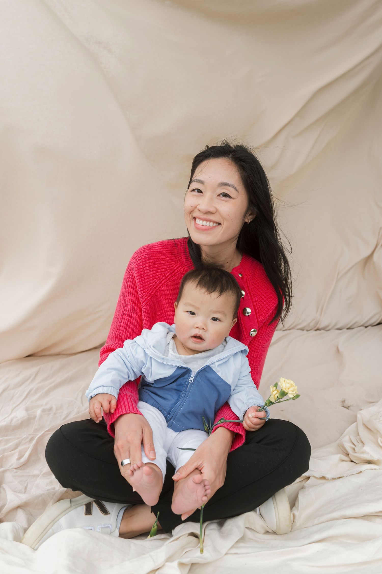 Smiling Asian woman in red cardigan sits cross-legged holding a baby in blue outfit, with a white flower, on beige fabric.