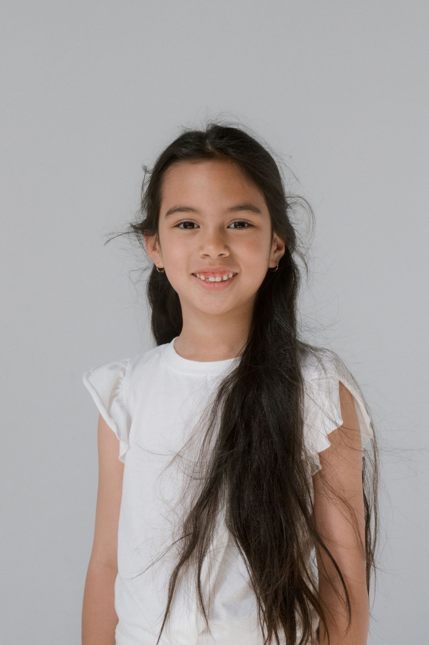 Smiling young girl with long dark hair wearing a white sleeveless top, posing against a light gray background.