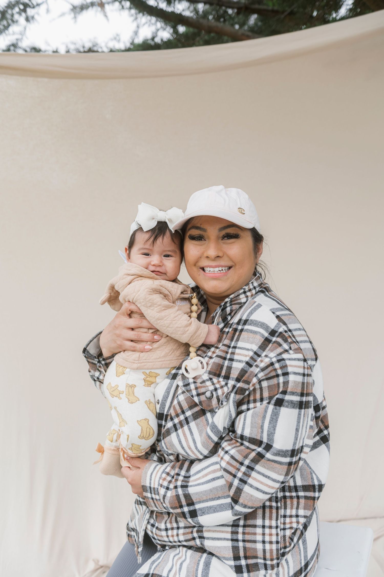 A smiling woman in a plaid jacket and white cap holds a baby girl with a bow outdoors.
