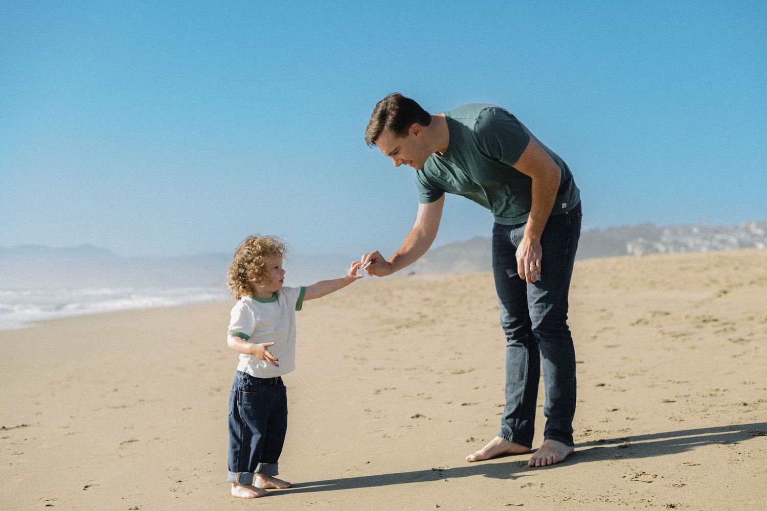A father holds his toddler's hand on a sunny sandy beach with clear blue skies and ocean waves in the background.