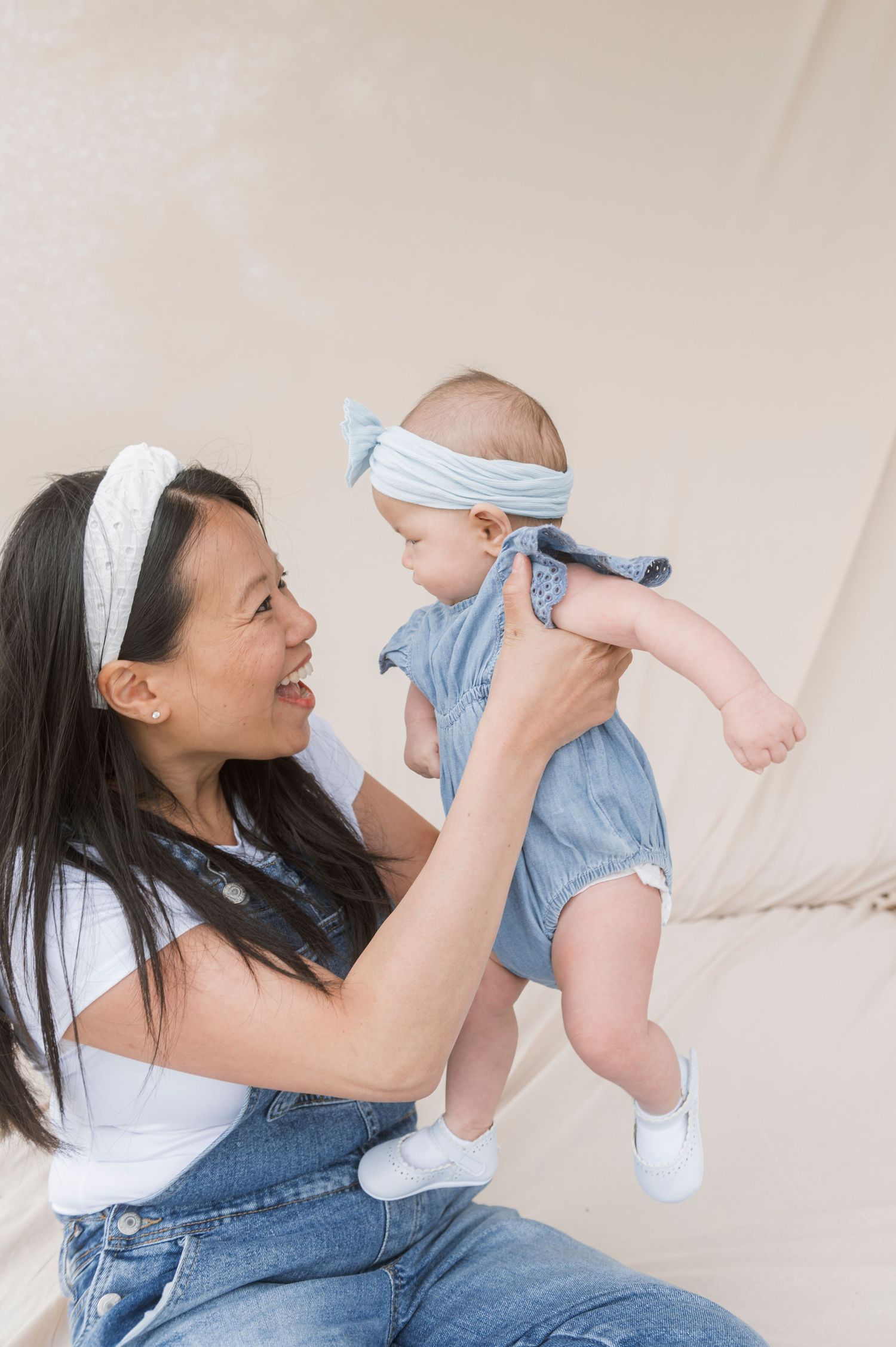 A smiling mother in denim overalls lifts her baby girl wearing a blue romper and headband in a bright studio.