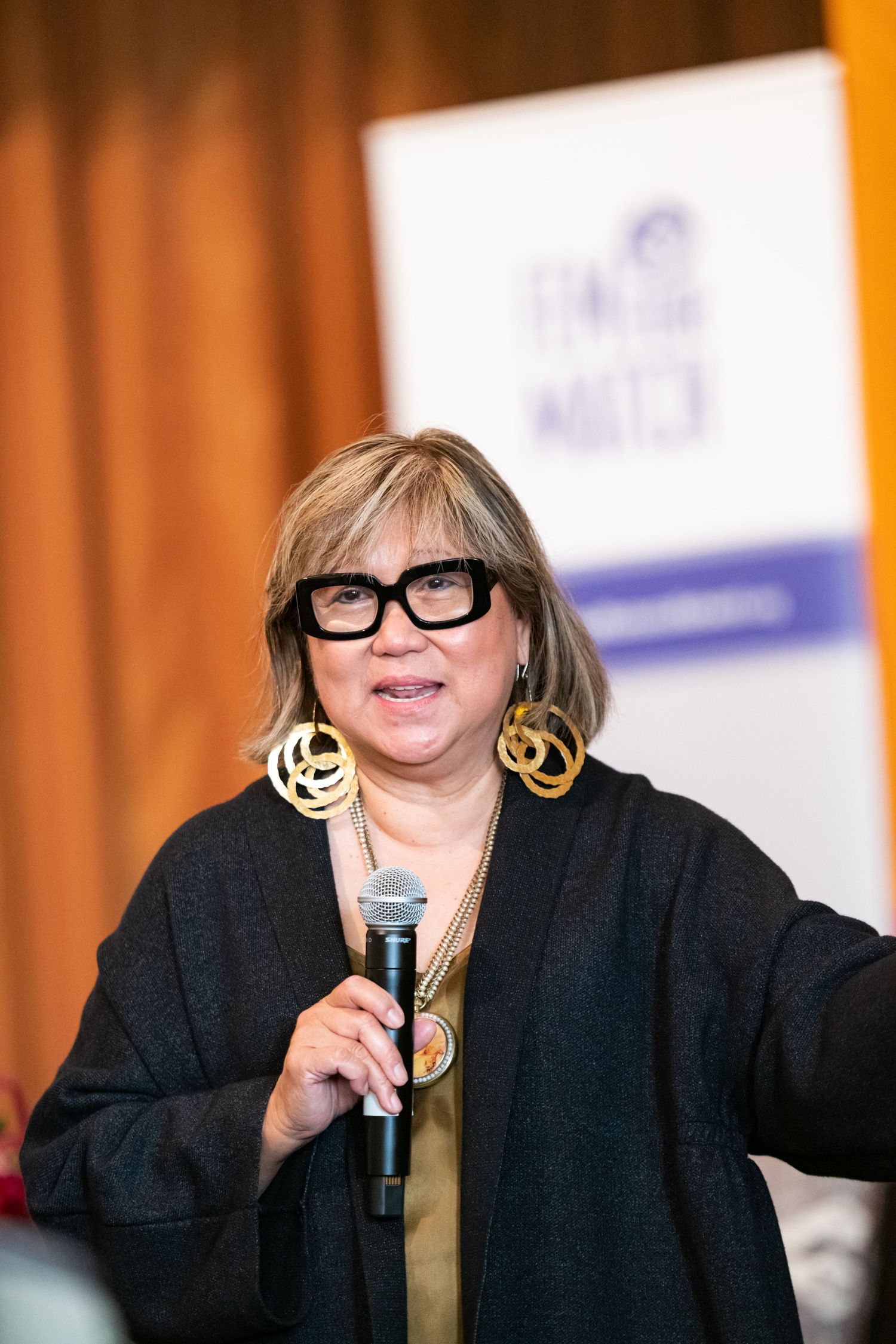 Woman with bold black glasses and gold earrings speaking into a microphone at an indoor event.