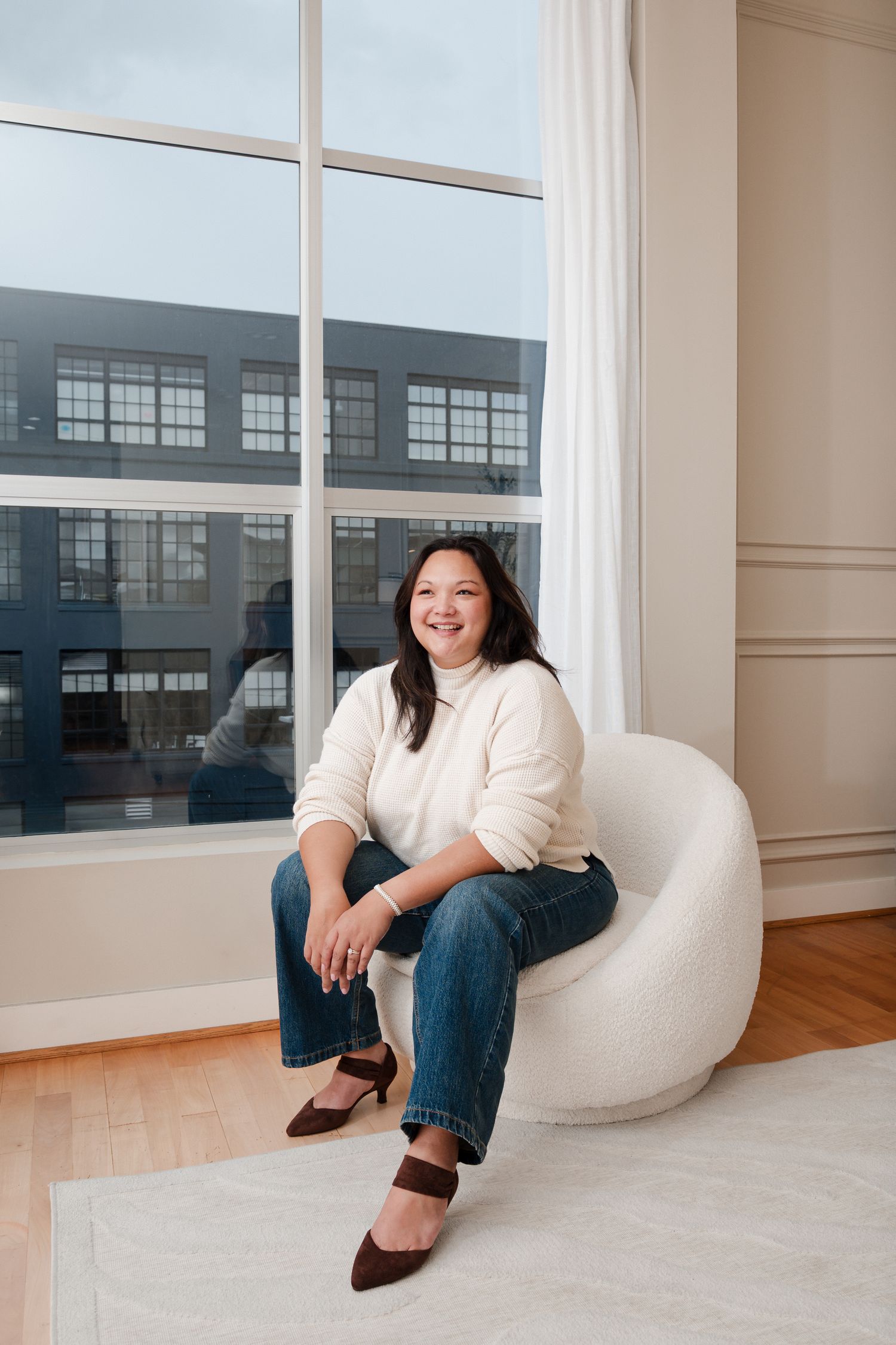 Woman in cream sweater and jeans sits smiling in white chair by large window in bright modern interior.