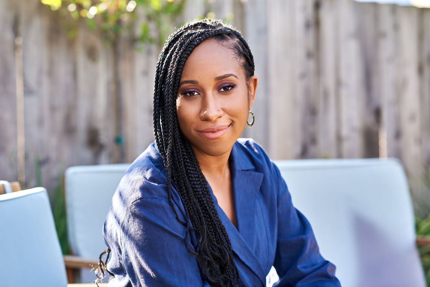 A woman with long box braids wearing a blue blazer smiles confidently while seated outdoors near a wooden fence.