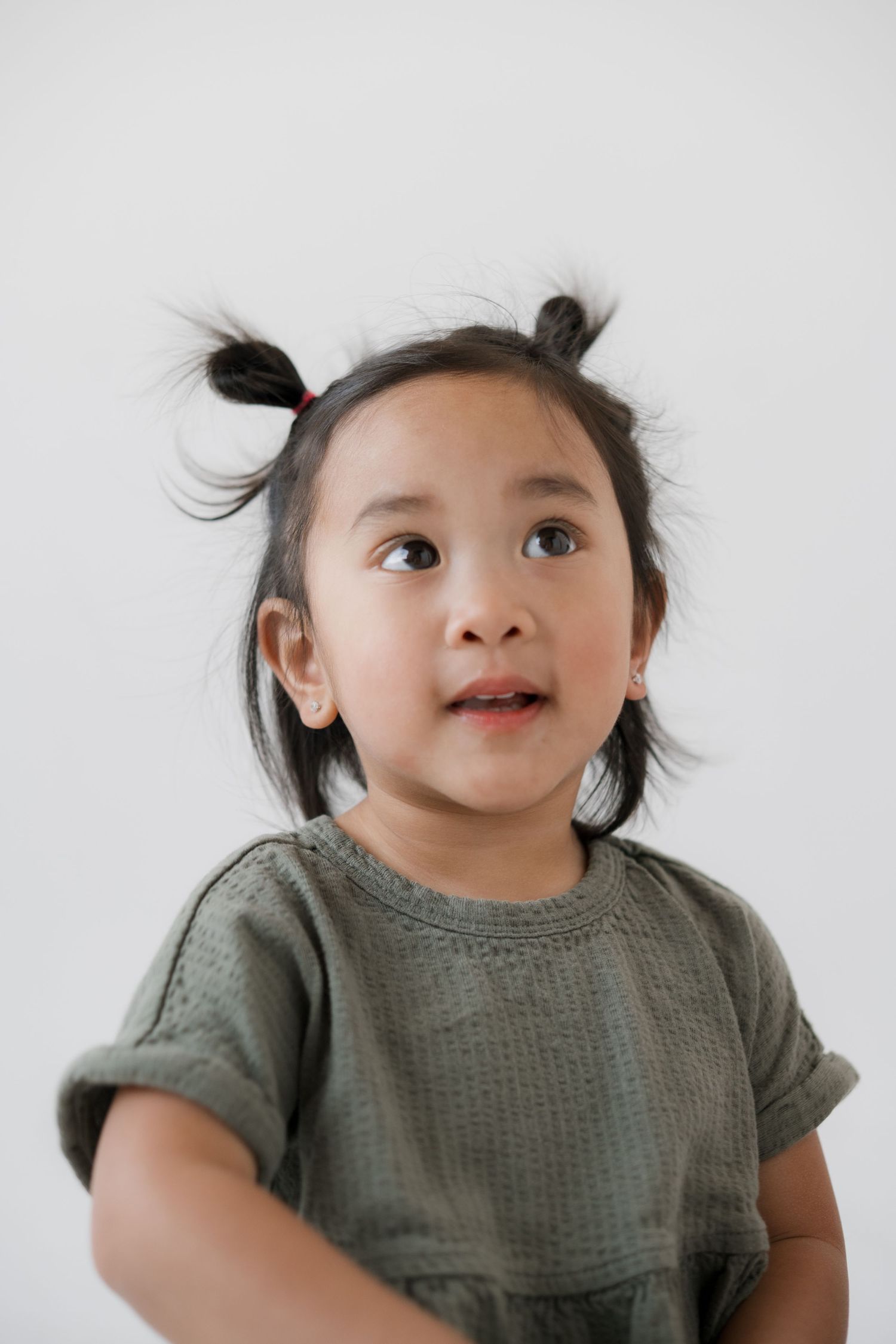 A cute young Asian girl with two small pigtails wearing a green top, looking up curiously against a white background.
