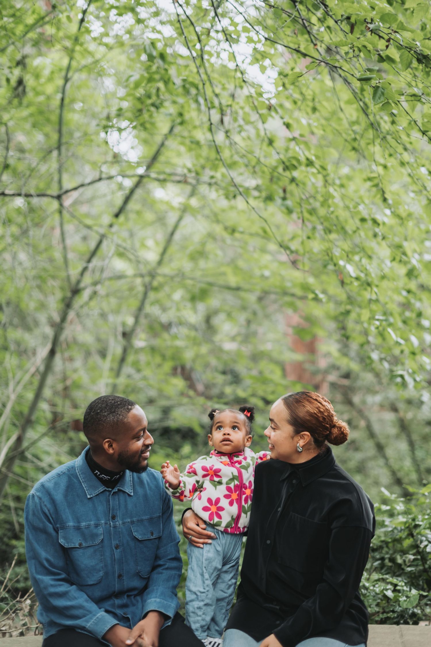 A young Black family with a baby girl sits outdoors surrounded by lush green trees in a park setting.