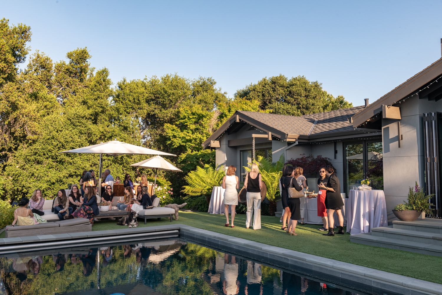 People gather in a backyard setting with umbrellas and outdoor furniture during a daytime event.