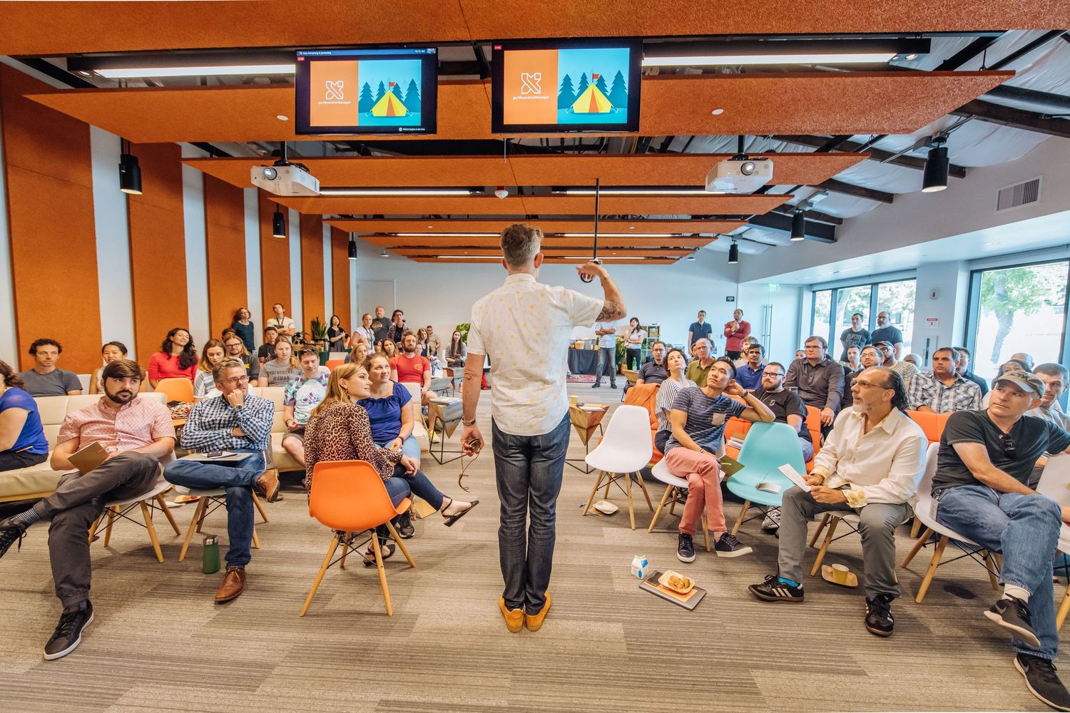 A panoramic view of people sitting in orange chairs during a meeting in a modern office space with large windows.