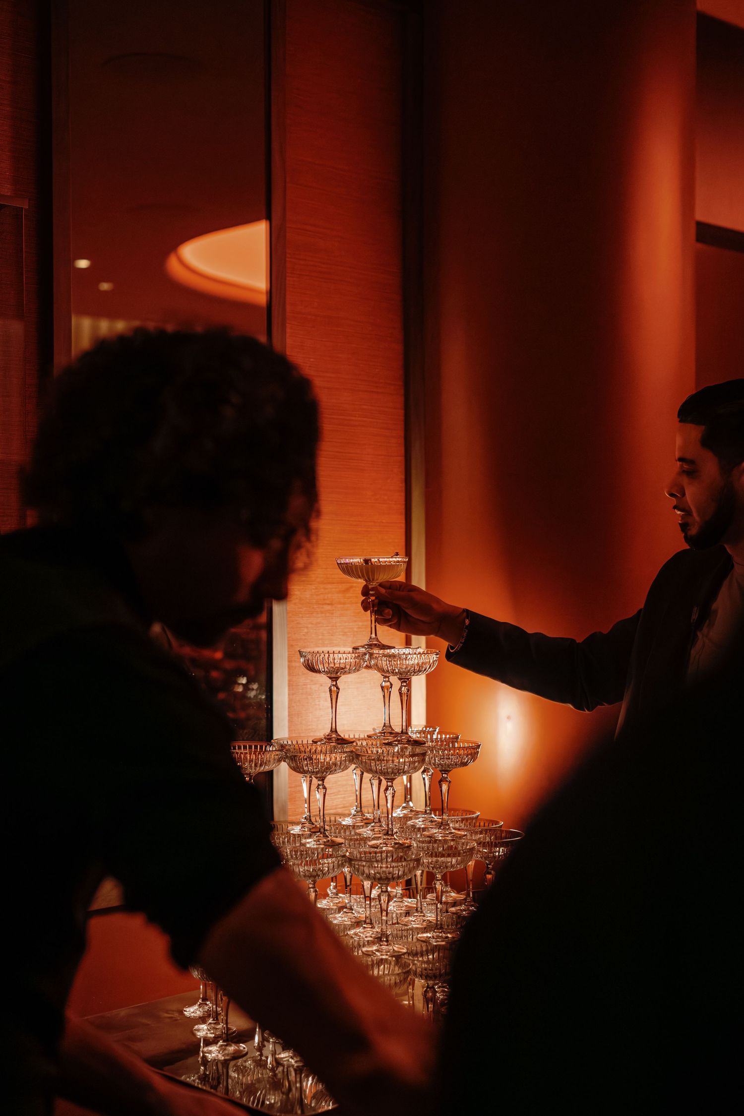 Silhouetted figures arrange champagne glasses in a tower formation under warm amber lighting at an elegant event.
