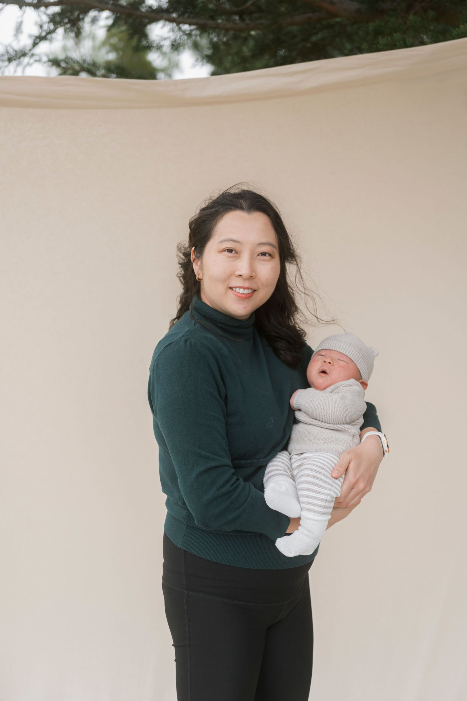Smiling woman in green turtleneck holding a newborn baby dressed in gray stripes outdoors.
