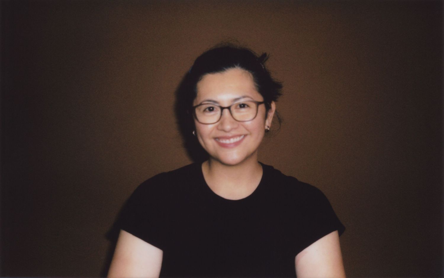 Smiling young person with glasses and short dark hair wearing a black t-shirt against a brown background.