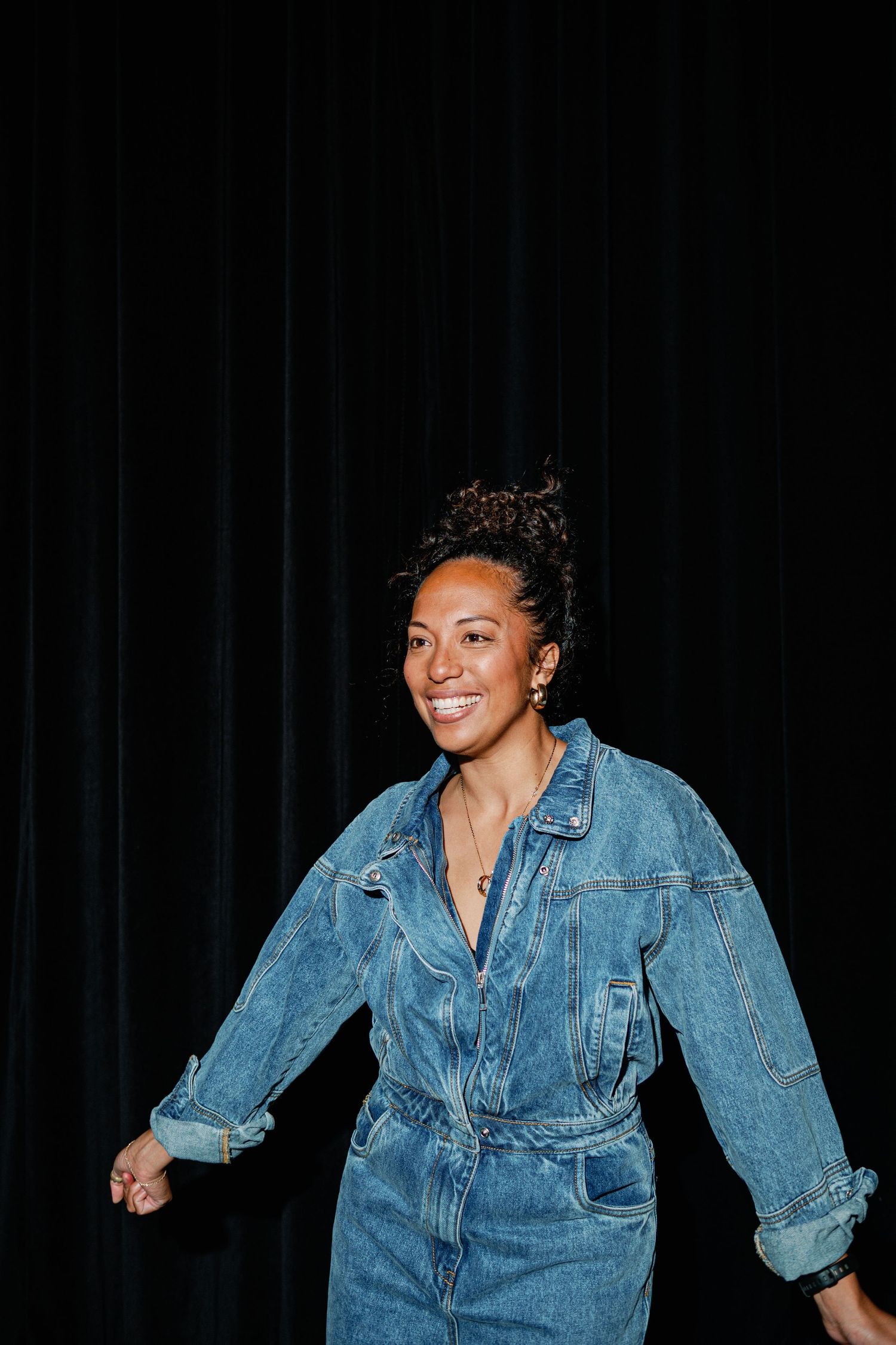 Smiling woman in denim jumpsuit posing against black curtain backdrop.