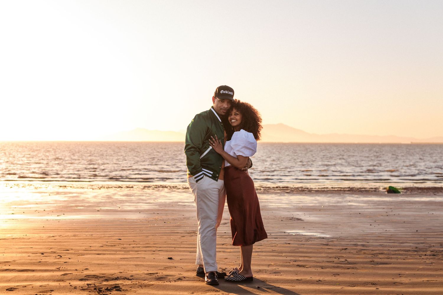 A couple embraces on a sandy beach at sunset creating a romantic silhouette against the orange sky.