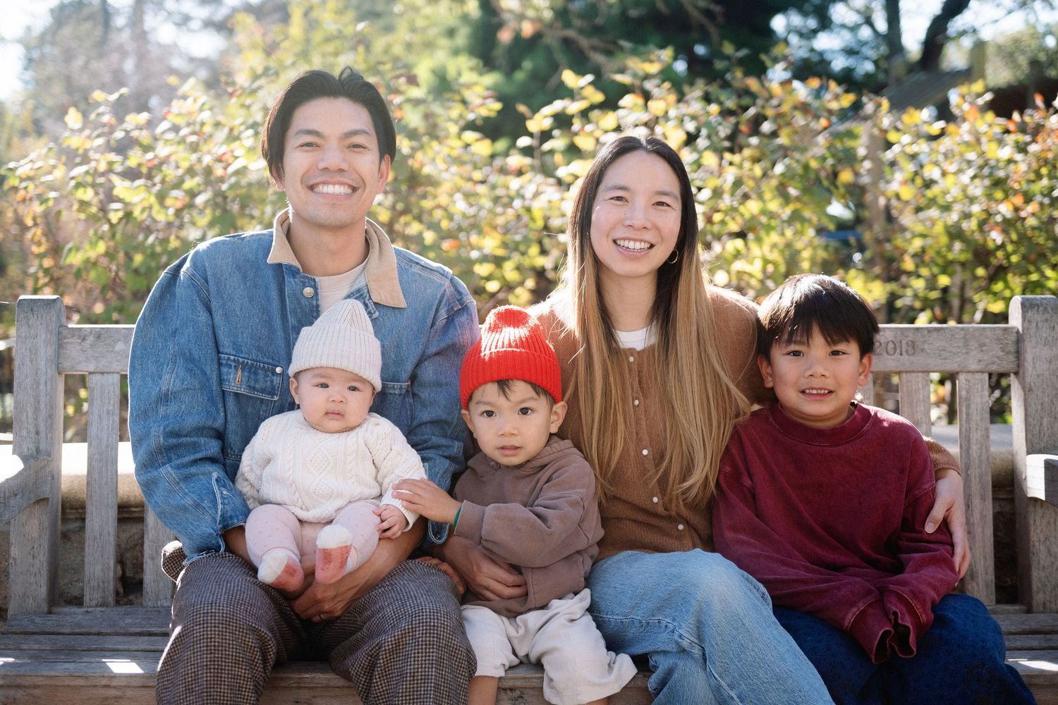 Happy Asian family with three children sitting on a wooden bench outdoors in a sunny autumn park.