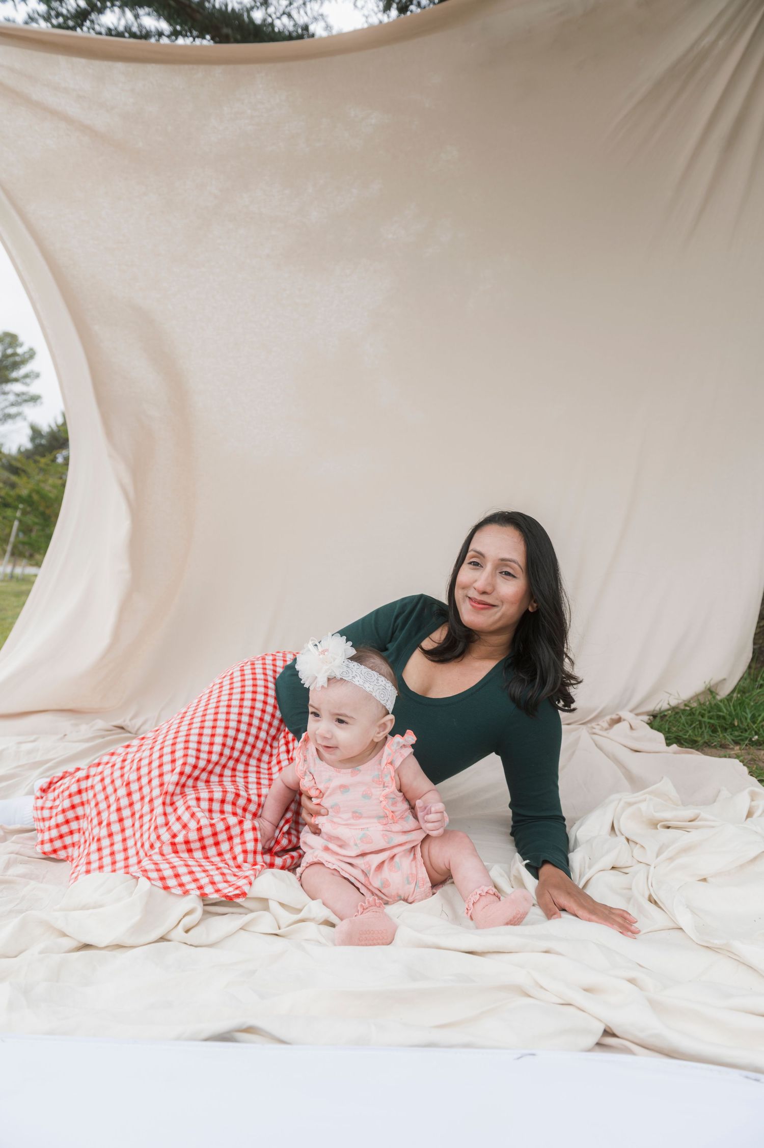 Mother and baby girl sitting together on cream fabric backdrop outdoors, baby wearing pink floral outfit with white bow headband.