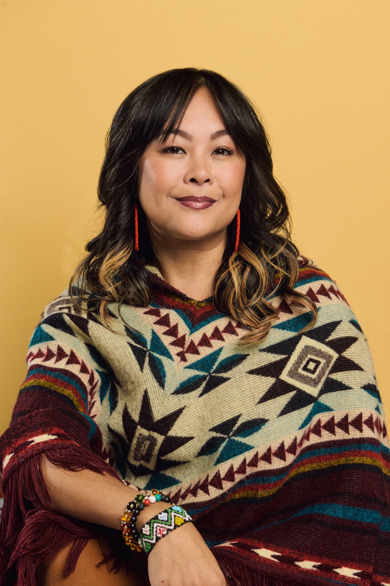 Woman in Native American geometric patterned poncho with red earrings and beaded bracelet posing against yellow background.