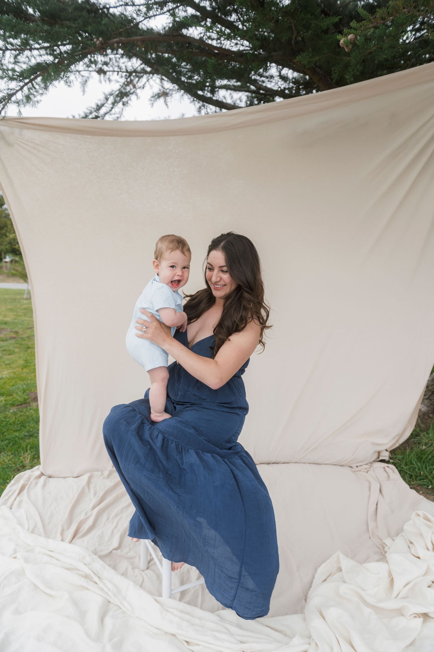 A woman in a navy blue dress holds a baby outdoors against a cream fabric backdrop during a professional photo session.