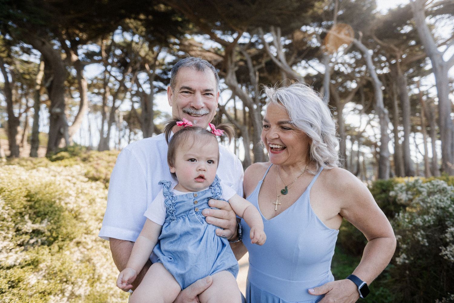 Smiling grandparents holding a baby girl in a blue dress outdoors with trees in the background.