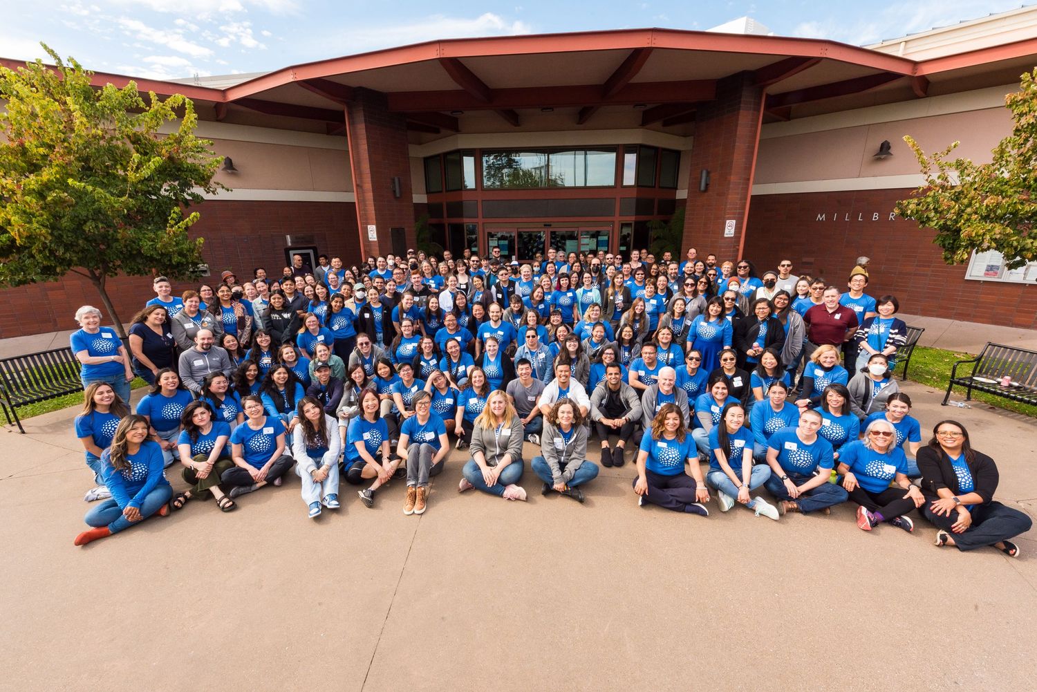Large group of students and staff in blue shirts posing outside a brick school building for a group photo.