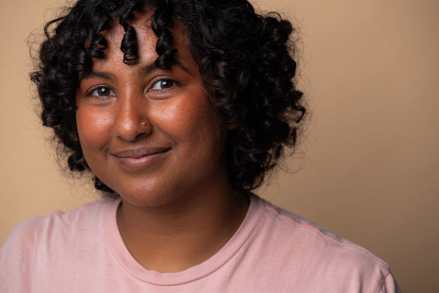 Young woman with curly black hair and light pink shirt smiling softly against a beige background.