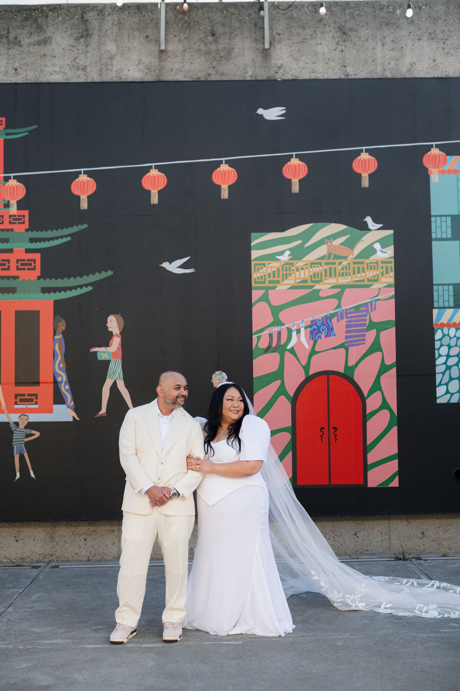 A wedding couple poses in front of a colorful Asian-inspired mural with red lanterns and decorative elements.
