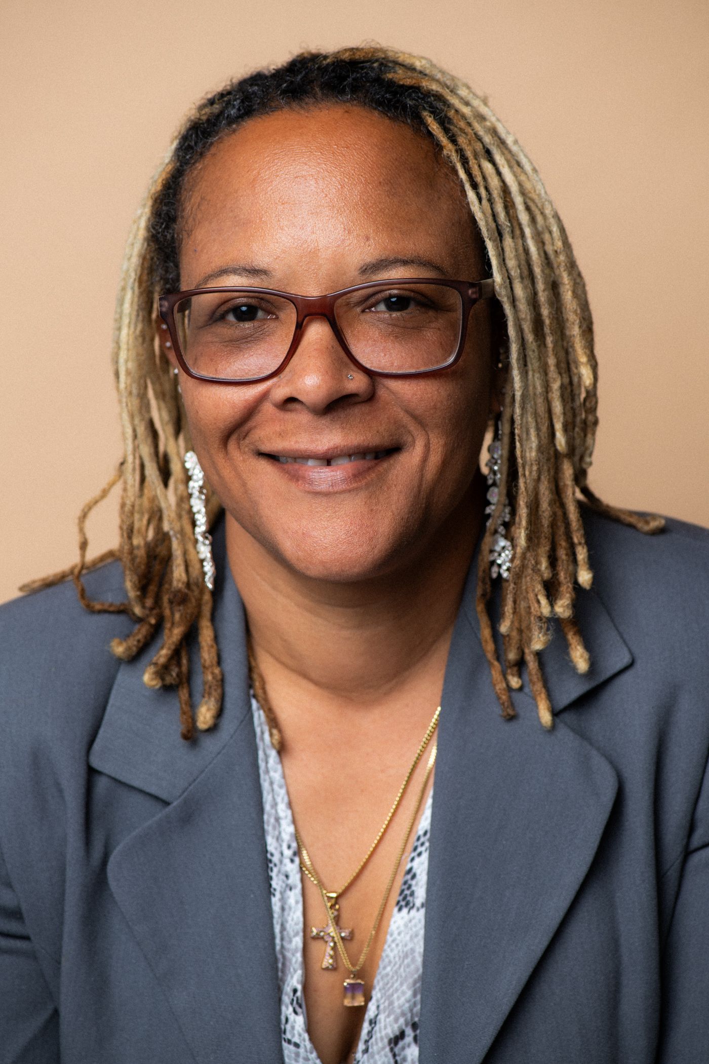 Professional headshot with gray blazer and blonde dreadlocks against neutral background.