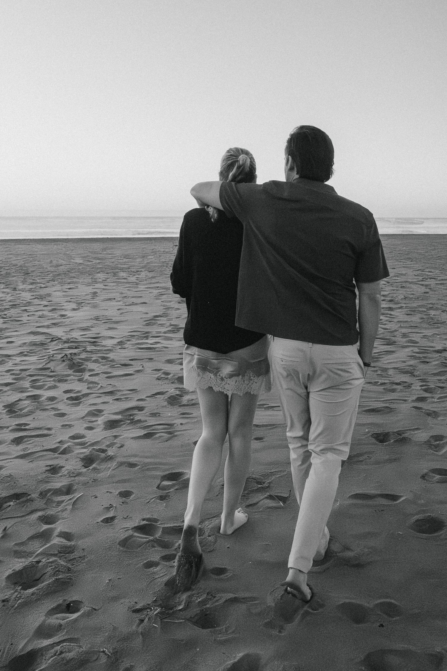 Black and white photo of a couple walking together on a beach with footprints in the sand.