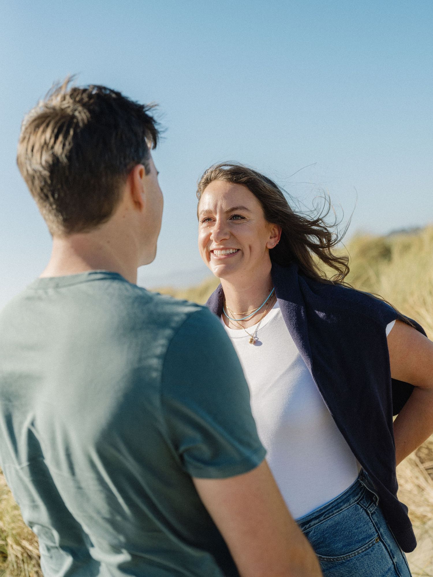 A smiling woman with windswept hair in a white top and navy vest chats with a man in a teal shirt outdoors.