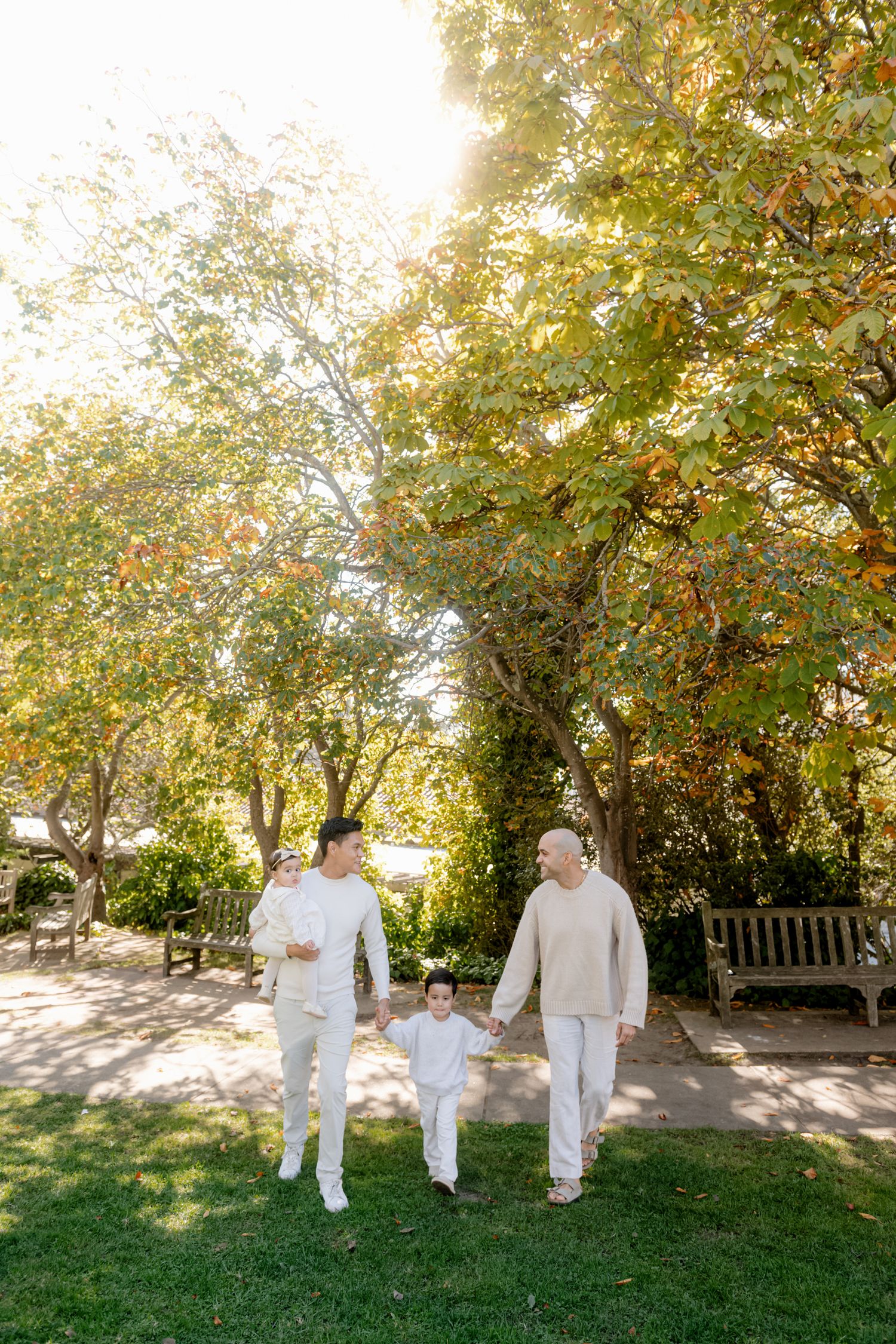 Three people in coordinated white outfits stroll through a garden setting.