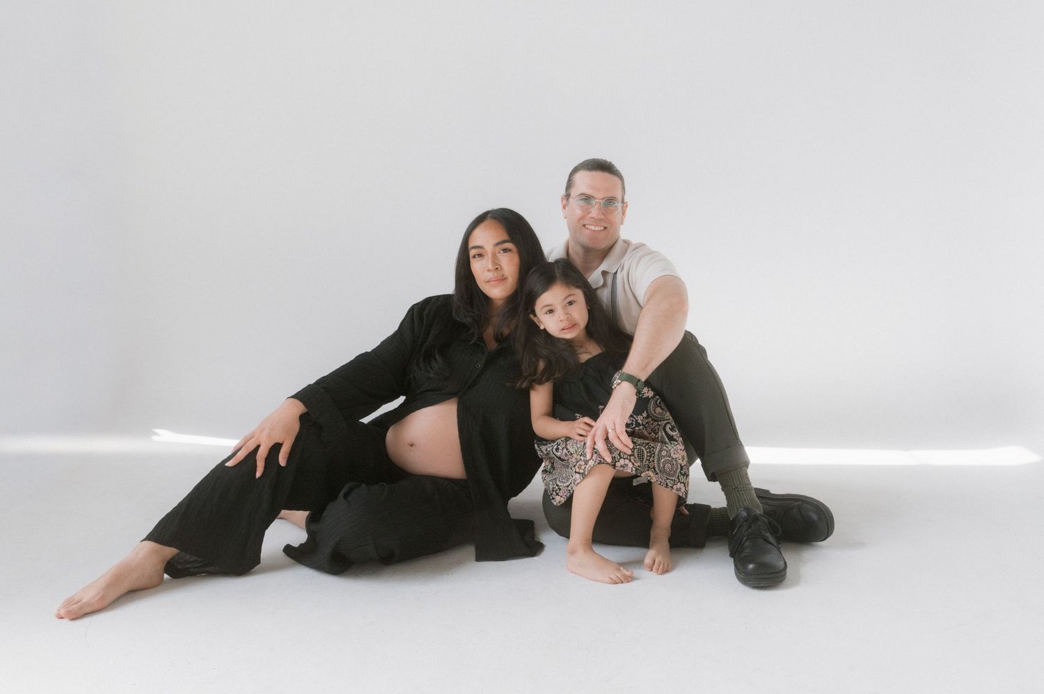 A family of three poses together on a white studio floor, with a pregnant woman, man, and young toddler girl.