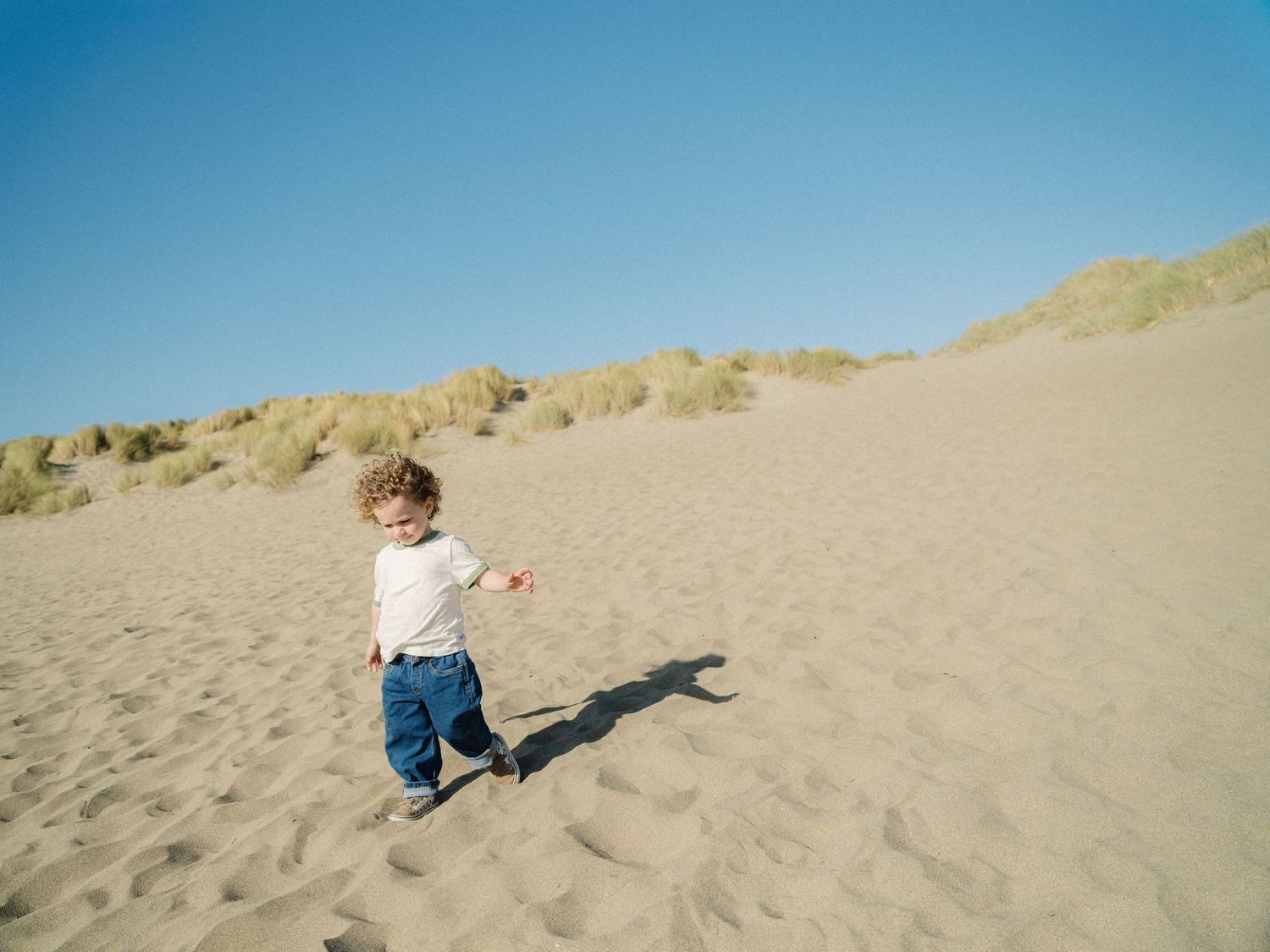 A toddler walks across sandy dunes with sparse grass under a clear blue sky.