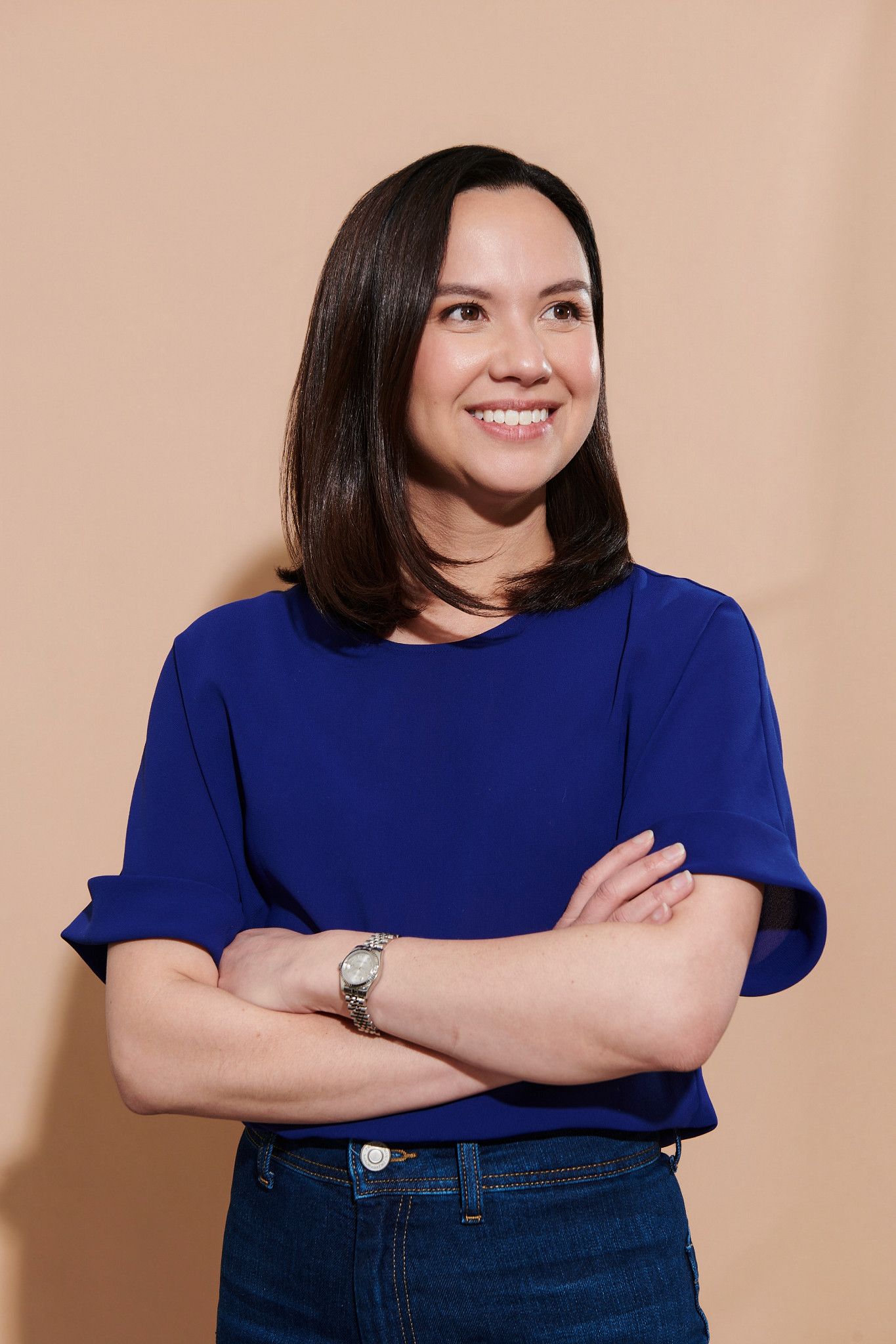 Smiling woman in blue top and jeans with arms crossed, posing confidently against a beige background.