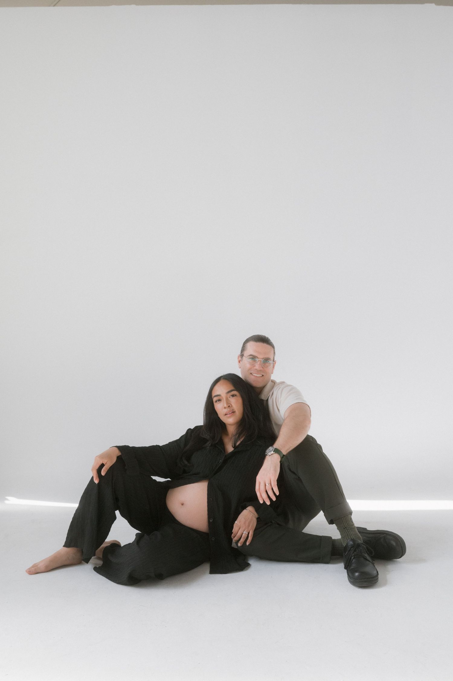 Pregnant couple sitting on white studio floor, woman in black suit showing baby bump, man in white shirt beside her.