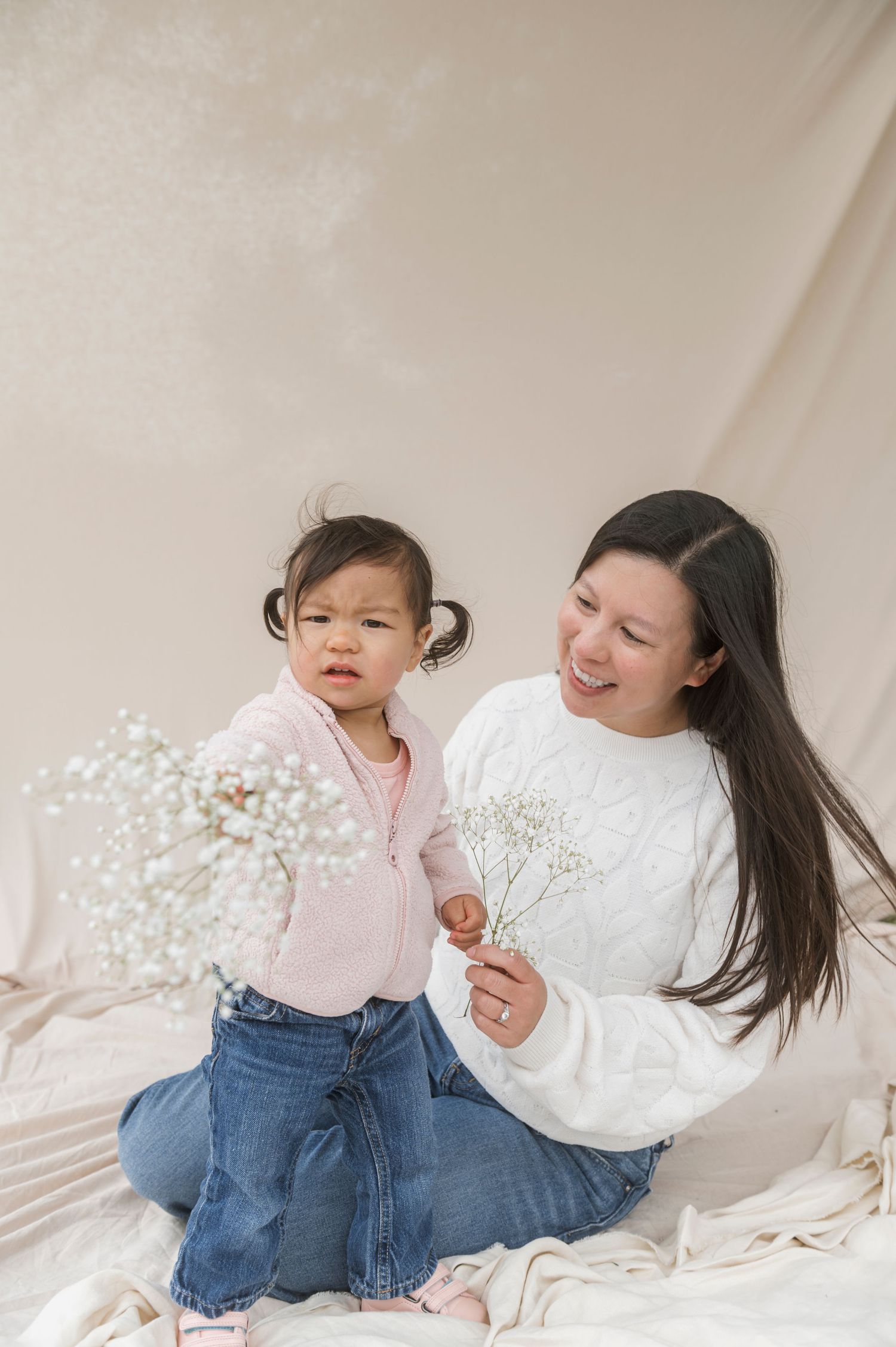 A woman and toddler girl sit together holding white baby's breath flowers against a neutral backdrop.