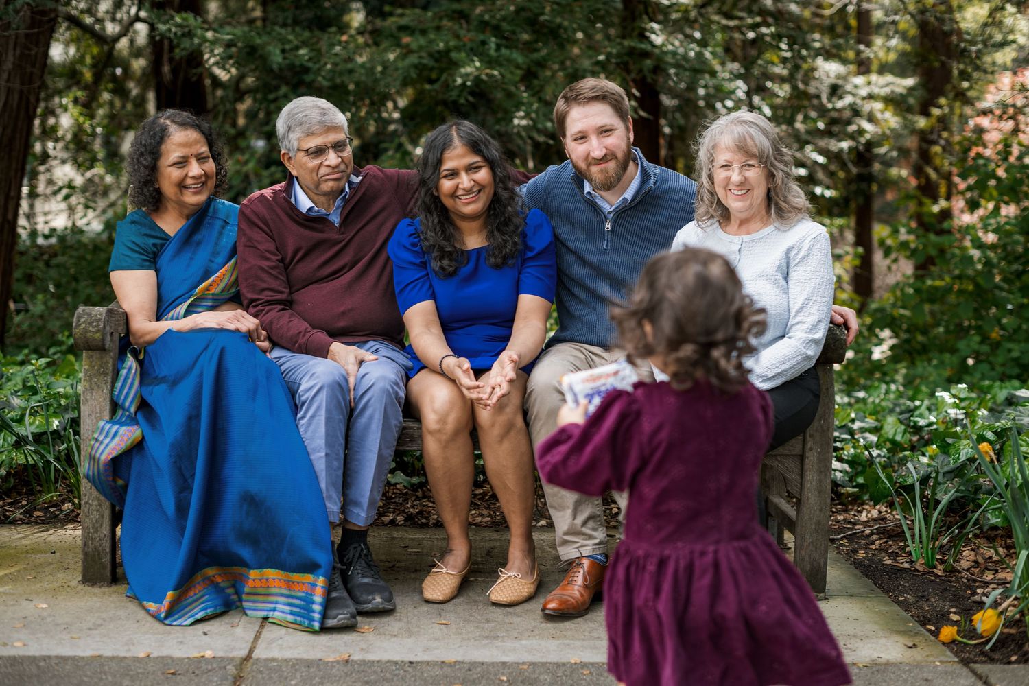 Multi-generational family smiling on a garden bench as a young child photographs them outdoors.