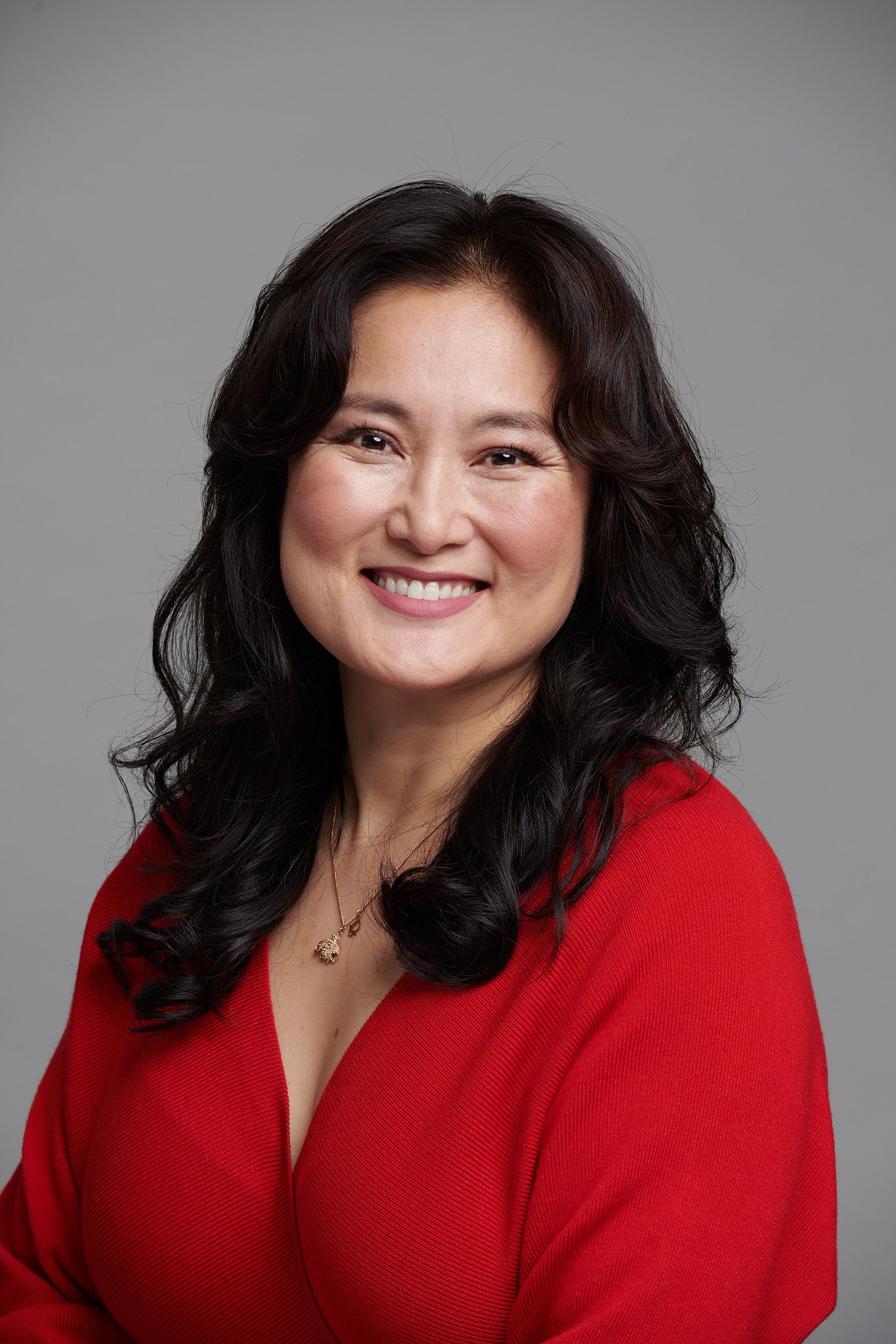 Professional headshot portrait of someone wearing a bright red sweater against a gray background, smiling warmly.