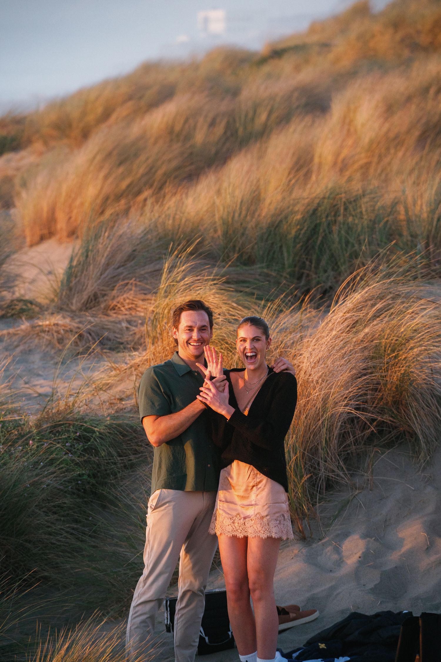 A romantic couple laughs together against a backdrop of beach grass at golden hour.