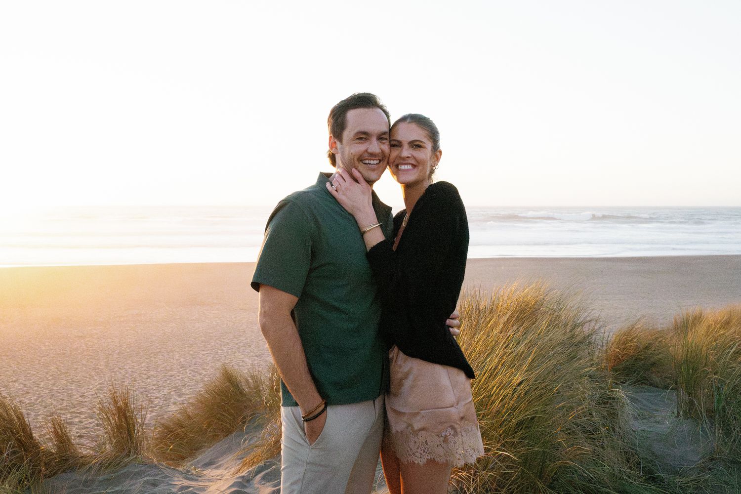A couple enjoys a tender moment on a scenic beach with dunes and ocean views.