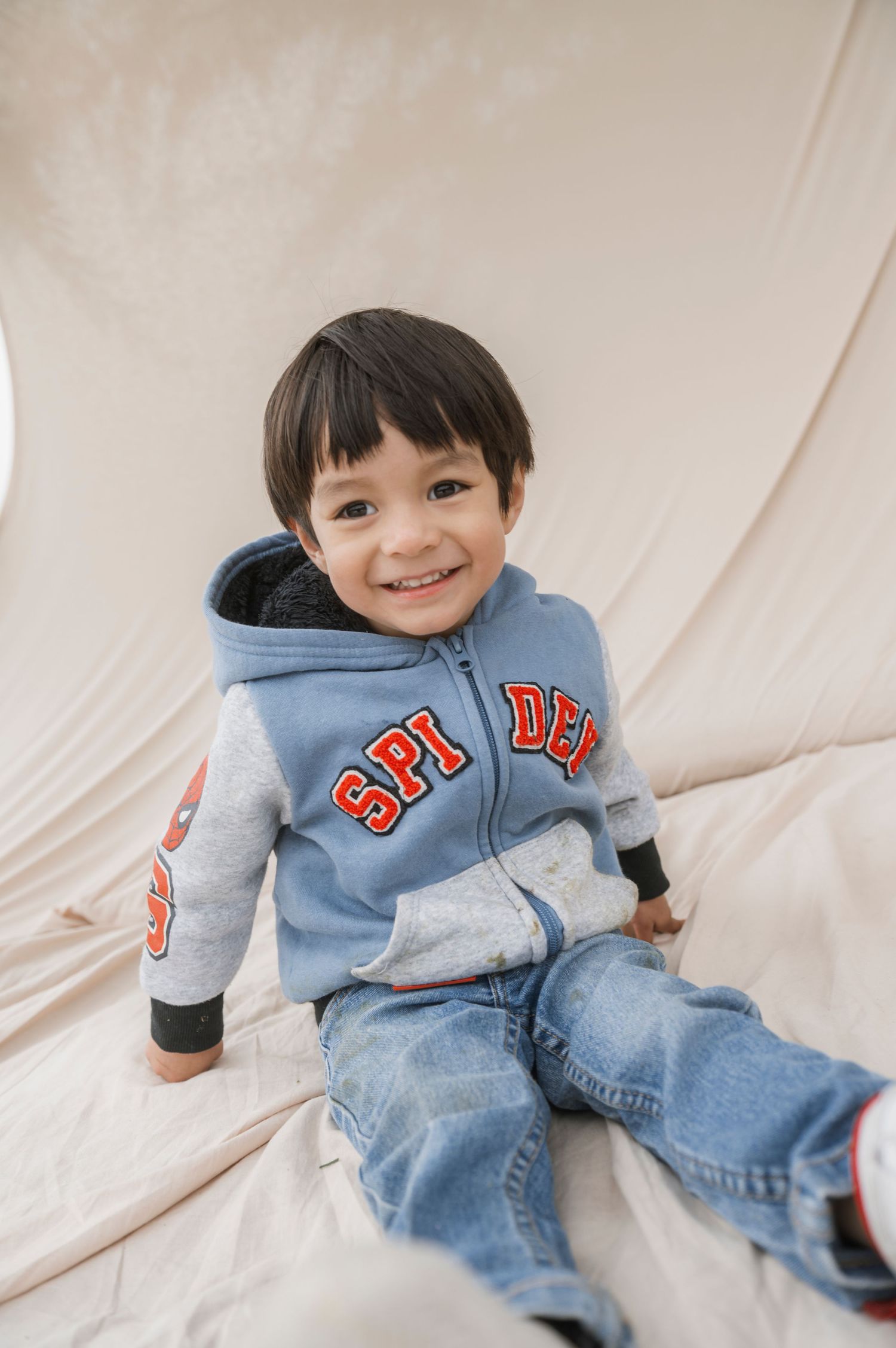 Smiling toddler boy wearing a Spider-Man zip-up hoodie and jeans sitting on a light beige fabric background.