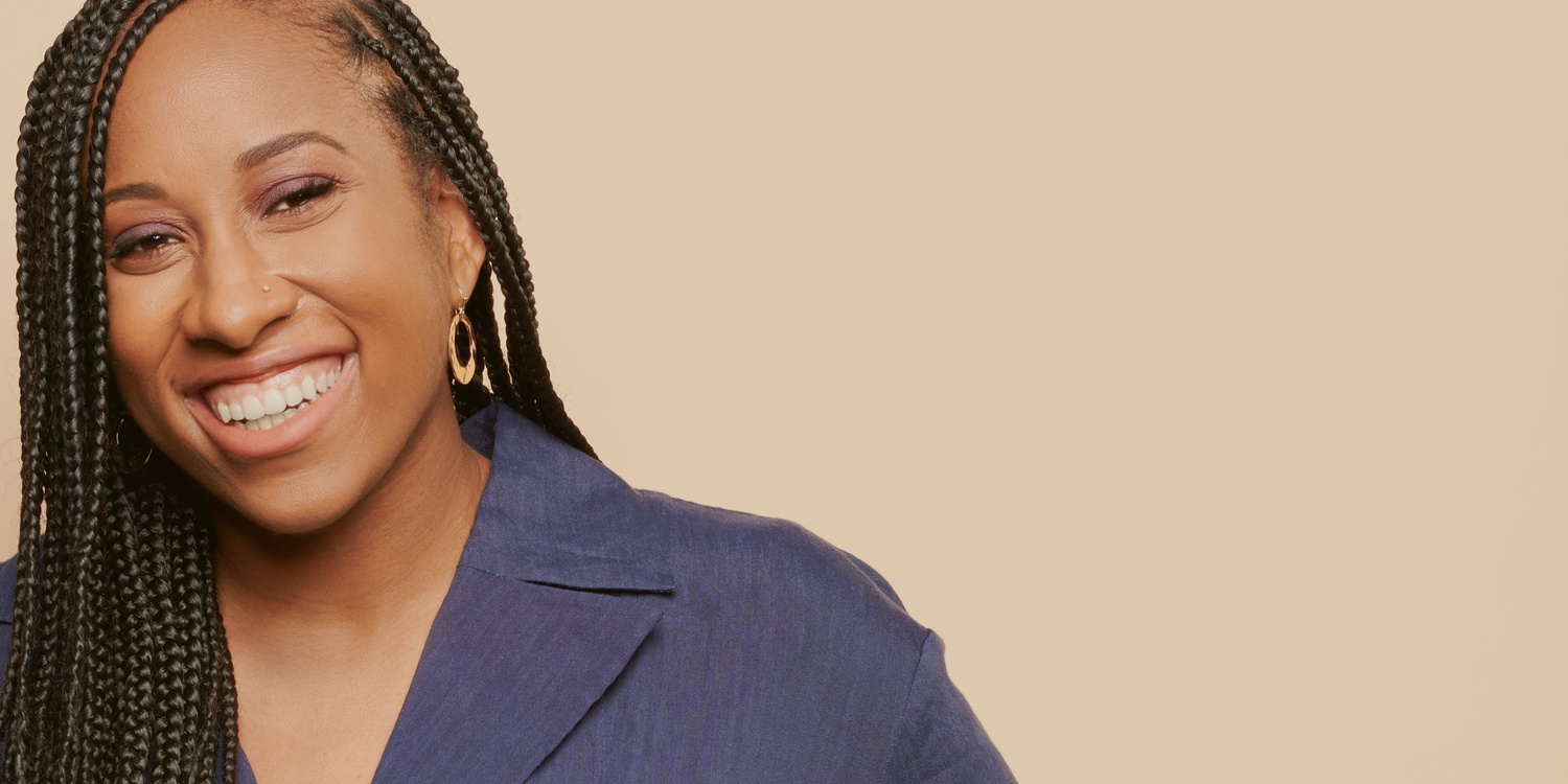 Smiling woman with long braids wearing gold hoop earrings and a blue blazer against a beige background.