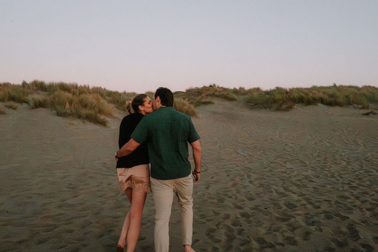 Two people walk hand in hand along an empty beach at sunset.