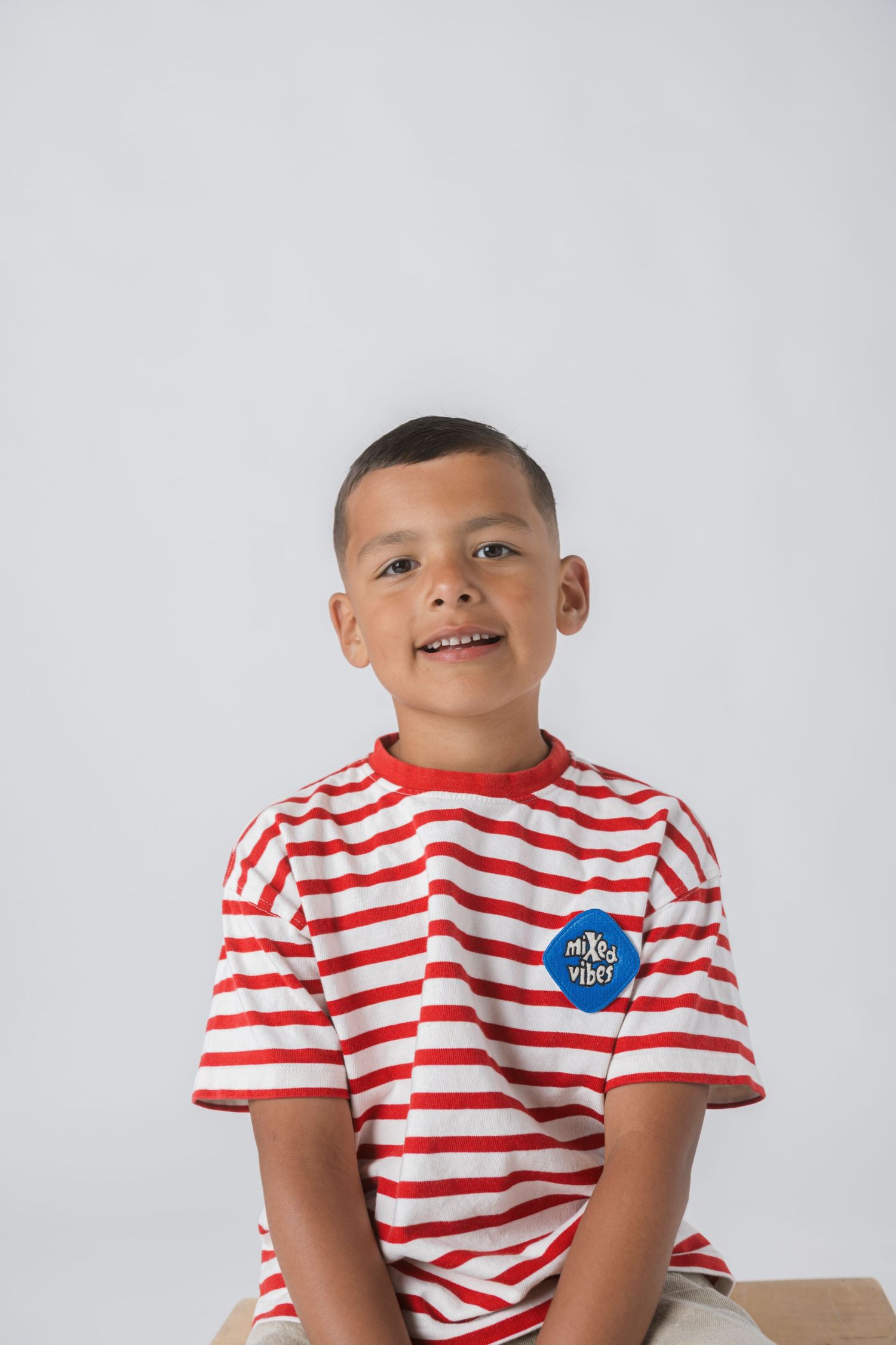 A smiling young boy wearing a red and white striped t-shirt with a blue badge, sitting against a light gray background.