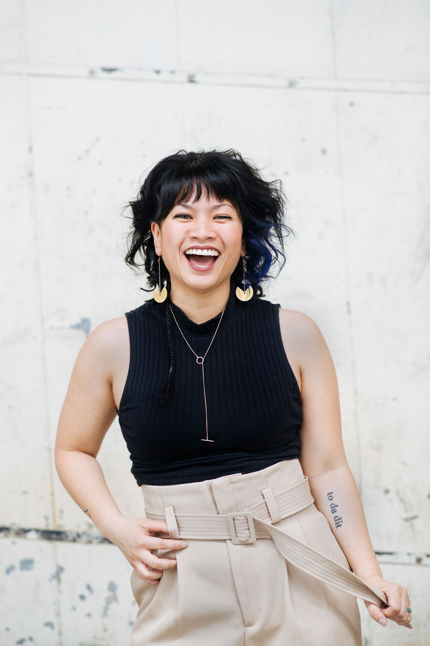 Laughing Asian woman with short black hair, gold earrings, black sleeveless top, and beige belted pants against a wall.