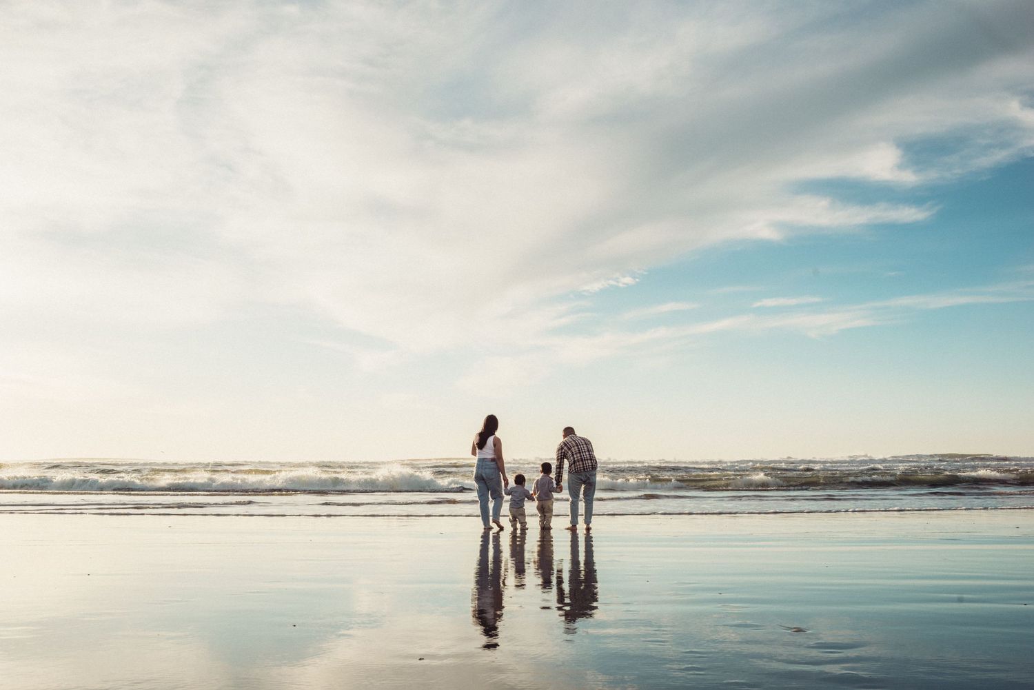 Family of four walking hand-in-hand on a reflective wet beach with ocean waves and blue sky.
